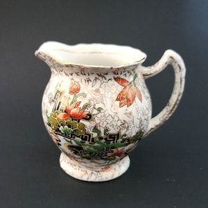 May include: A small, vintage cream pitcher with a floral design. The pitcher is white with gold trim and features a colorful pattern of flowers and foliage in shades of red, green, and orange. It has a curved handle and a small spout.