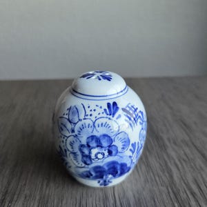 May include: A small, round, lidded ceramic jar with a blue and white floral design. The jar features a detailed pattern of blue flowers and leaves against a white background. The lid has a small floral design on top.