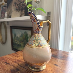 May include: An ornate, antique-style pitcher with a beige body and intricate gold, blue, and red patterns. The pitcher has a gold handle and trim, and sits on a wooden table. The design is reminiscent of vintage decor.