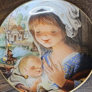 May include: Decorative plate featuring a serene illustration of a woman and a baby, with a village scene in the background. The plate has a gold rim and the artist's signature. The woman wears a white head covering and a blue dress.