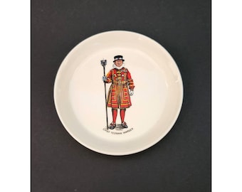 May include: A white ceramic dish featuring a detailed illustration of a Yeoman Warder in a red and gold uniform, holding a staff. The text "CHIEF YEOMAN WARDER" is printed at the bottom of the image. The dish has a simple, round shape.