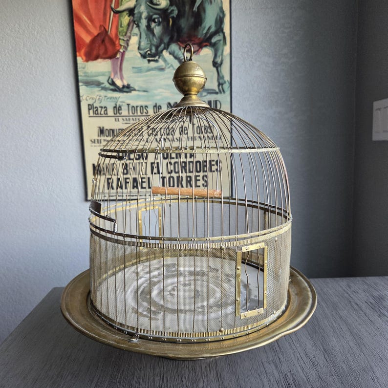 May include: An antique brass birdcage with a domed top and a circular base. The cage features a mesh screen and a small door. A vintage poster is in the background.