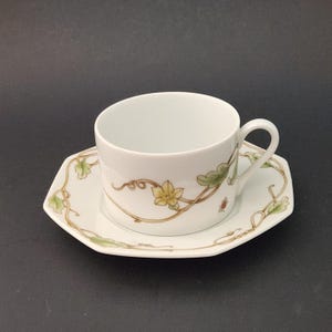 Op de afbeelding: Witte theekop en schotel set. De kop en schotel zijn versierd met een delicaat bloemmotief in groen, geel en bruin. De schotel heeft een achthoekige vorm. Een klein rood lieveheersbeestje is ook zichtbaar.