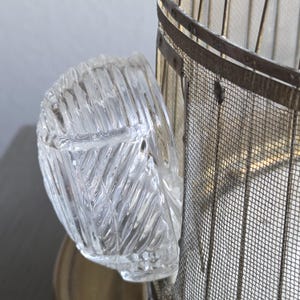 May include: A clear, textured glass container with a curved shape, possibly a small bowl or ashtray. The glass has a repeating diagonal pattern. The container is positioned next to a metal mesh structure.