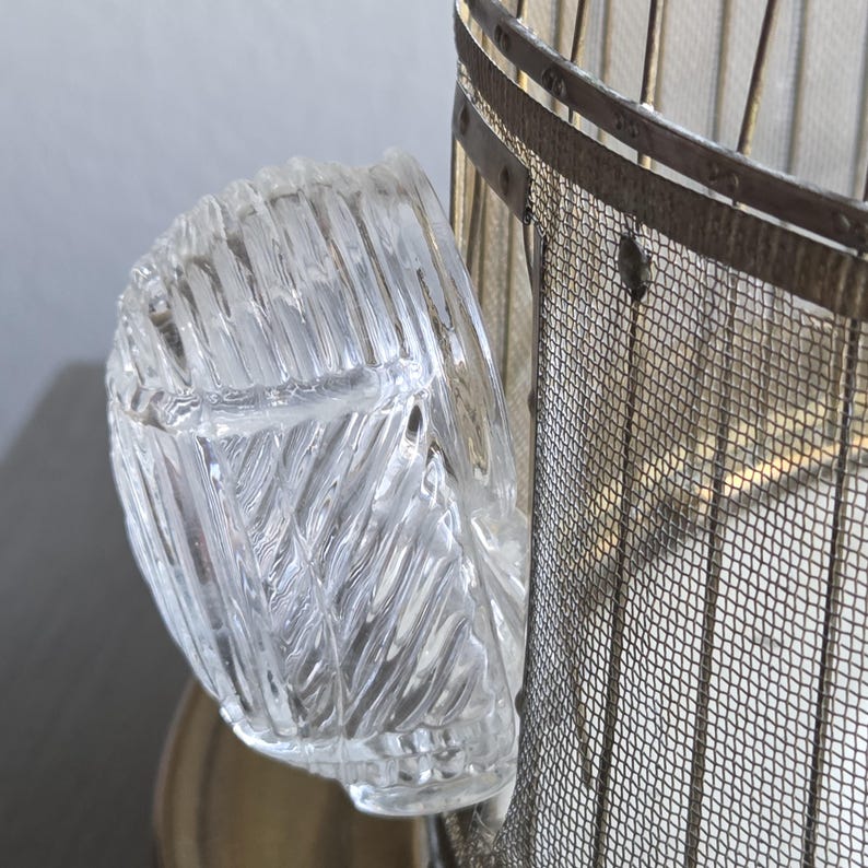 May include: A clear, textured glass container with a curved shape, possibly a small bowl or ashtray. The glass has a repeating diagonal pattern. The container is positioned next to a metal mesh structure.