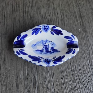 May include: A small, white ceramic ashtray with a blue floral and windmill design. The ashtray has a scalloped edge and two indentations for resting cigarettes. The interior features a blue landscape scene.
