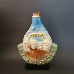 May include: A ceramic bottle with a cork stopper, featuring a painted depiction of Mount Rushmore and the text "BLACK HILLS". The bottle has a blue top and a green base with the words "MT. RUSHMORE".