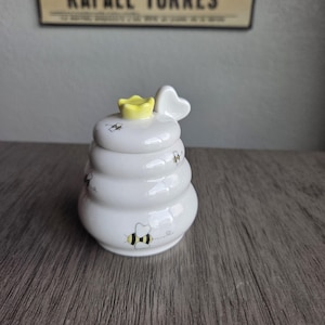 May include: A white ceramic honey pot shaped like a beehive, with a yellow flower on top and a heart-shaped handle. The pot is decorated with small black and yellow bee illustrations. The honey pot is a charming addition to any kitchen.