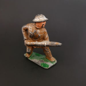 May include: A vintage toy soldier figurine in a brown uniform and helmet, holding a long, cylindrical object. The figure stands on a green and gray base. The paint shows signs of wear, suggesting its age.