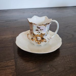 Könnte beinhalten: Ein weißes Tassen- und Untertassen-Set. Die Tasse hat einen gewellten Rand und einen Henkel. Sie ist mit goldenen floralen Designs verziert. Die Untertasse ist ebenfalls weiß und hat einen gewellten Rand.
