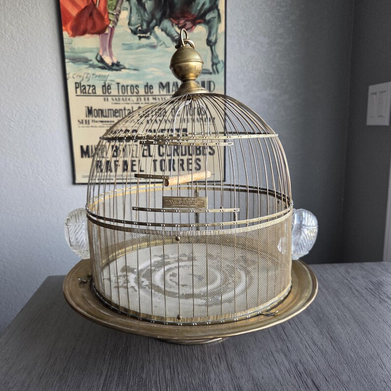 May include: Vintage brass birdcage with a domed top, mesh sides, and a circular base. The cage features a small perch and glass handles. A decorative piece for home decor.