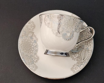 May include: A white teacup and saucer set with a silver floral design. The cup has a silver handle and base, and the saucer has a matching pattern. The set is on a black background.