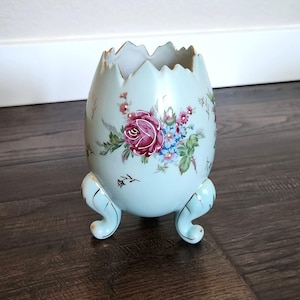 Puede incluir: Un jarrón de cerámica azul claro en forma de huevo con un diseño floral y detalles dorados. El jarrón tiene tres patas doradas.