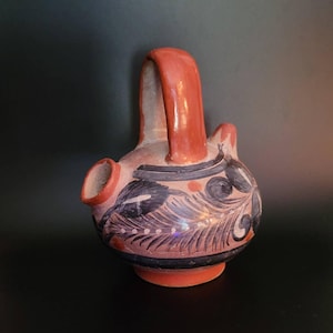 May include: A hand-painted ceramic pitcher with a red handle and spout. The body of the pitcher is decorated with black and white floral patterns and red accents. The base and handle are a solid red color.