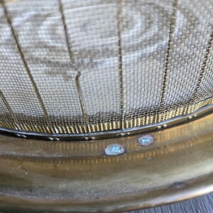 May include: Close-up of a vintage brass fire screen. The screen features a fine mesh design with a brass frame and rivets. The brass has a warm, aged patina with some areas of blue-green oxidation.