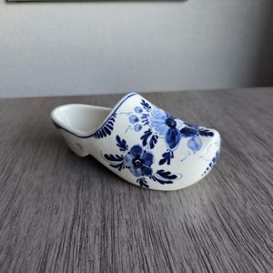 May include: A white ceramic Dutch clog, decorated with blue floral patterns. The miniature shoe features a rounded toe and a hollowed-out interior. The blue floral design includes stylized flowers and leaves, creating a classic, decorative piece.