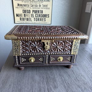 May include: A dark wooden jewelry box with intricate carvings and brass accents. The box features a hinged lid, decorative brass corners, and small drawers. The box has a vintage aesthetic, with a poster in the background that reads "Plaza de Toros de MADRID".