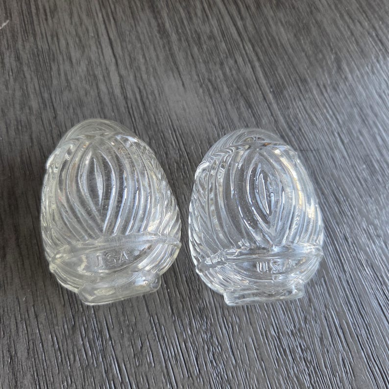 May include: Two clear, egg-shaped glass light fixture covers with a textured, swirled design. The base of each cover has the text "USA" etched into the glass. The covers are displayed on a gray wooden surface.