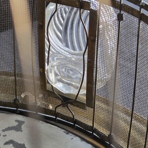 May include: Close-up of a clear glass lens with a spiral design, framed by a metal structure and wire mesh. The lens is encased in a rectangular frame with curved metal supports, suggesting a vintage or industrial aesthetic.