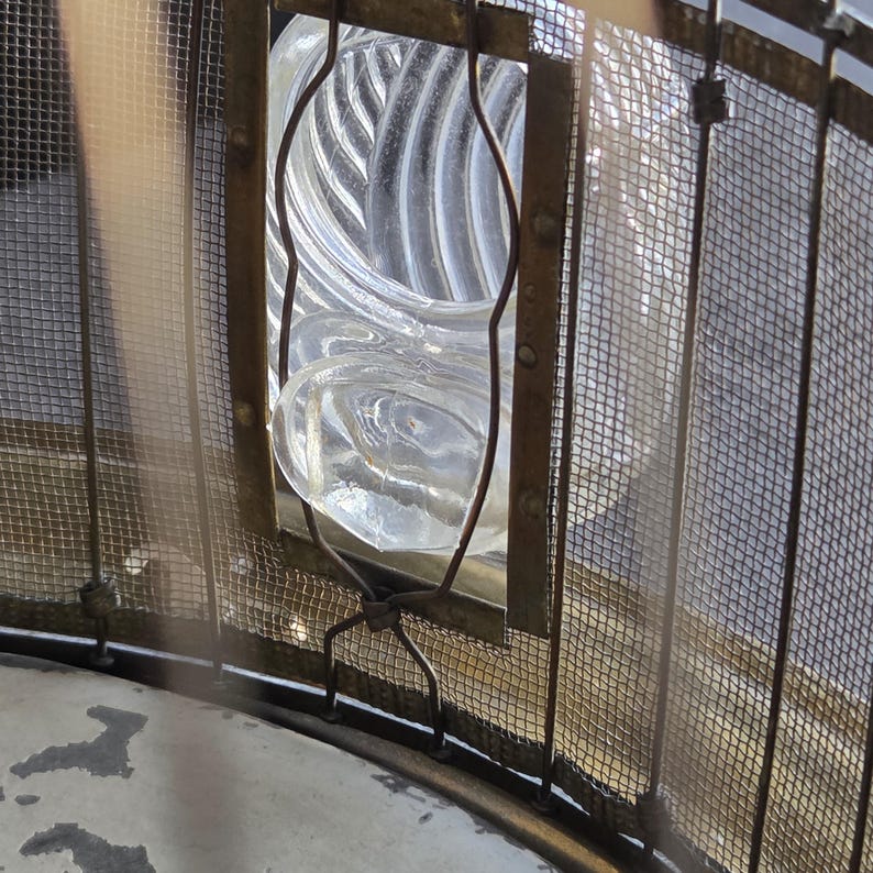 May include: Close-up of a clear glass lens with a spiral design, framed by a metal structure and wire mesh. The lens is encased in a rectangular frame with curved metal supports, suggesting a vintage or industrial aesthetic.