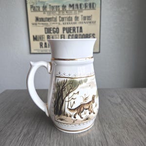 May include: White ceramic beer stein with a handle, featuring a gold rim and a painted scene of a dog in a field with trees. The stein has the text "Plaza de Toros de MADRID" on the background.
