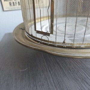 May include: An antique brass-toned food cover with a mesh screen and a circular base. The cover has a cage-like design with a hinged door and is likely used to protect food from insects. The base is wide and slightly curved.