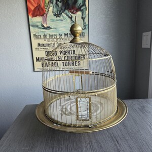May include: A vintage brass birdcage with a domed top and a circular base. The cage features a wire mesh design and a small door. A vintage poster is in the background.