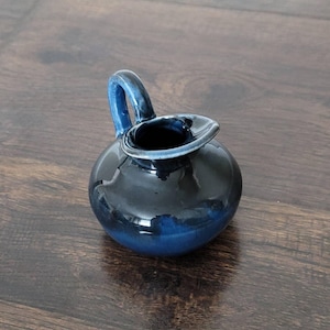 May include: A small, round, ceramic pitcher with a dark blue glaze. The pitcher has a curved handle and a spout. The color transitions from a deep navy to a lighter blue at the base. The pitcher is likely used for serving liquids.