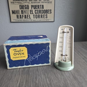 以下が含まれることがあります： ヴィンテージのテイラーオーブン温度計とレシピボックス。温度計はオフホワイトで、温度計と緑色のベースが付いています。レシピボックスは青色で、「Taylor Oven Thermometer Recipes」と書かれています。