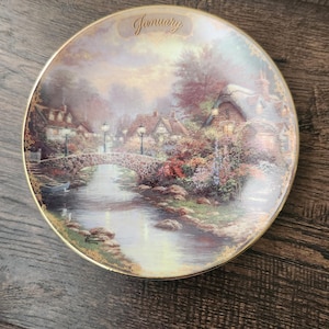 May include: Decorative plate featuring a scenic village landscape with a stone bridge over a river. The plate has a gold rim and the word "January" at the top. The artwork depicts a quaint village scene with houses and trees.