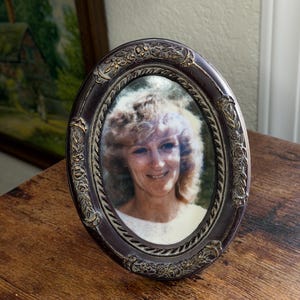 May include: Oval-shaped, dark brown picture frame with ornate floral detailing. The frame holds a portrait of a woman with light hair and a white top. The frame is displayed on a wooden surface.