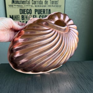 May include: A copper-colored, nautilus-shaped mold with a spiral design. The mold has a hanging loop and is likely used for baking or creating decorative desserts. The background includes a vintage poster with Spanish text.
