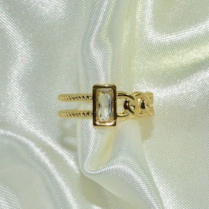 May include: A gold ring with a clear rectangular stone set in a gold frame. The ring has a chain link design on one side and a twisted rope design on the other.