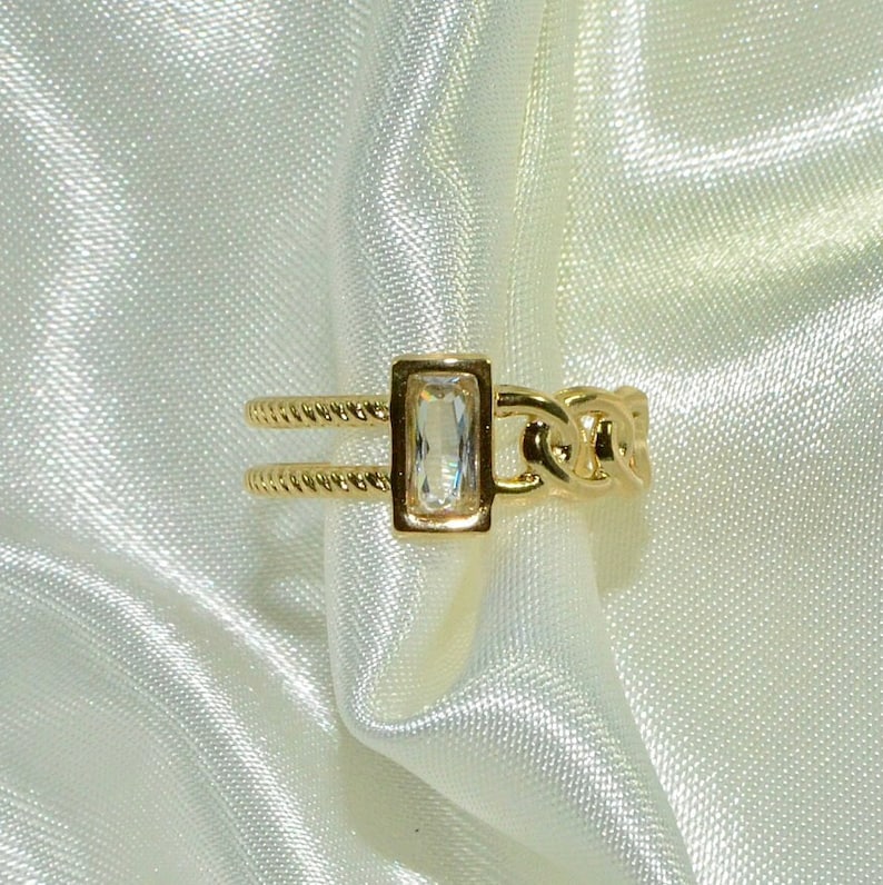 May include: A gold ring with a clear rectangular stone set in a gold frame. The ring has a chain link design on one side and a twisted rope design on the other.