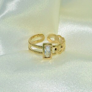 May include: Gold ring with a clear rectangular stone set in a gold frame. The ring has a chain-link design on one side and a twisted rope design on the other.