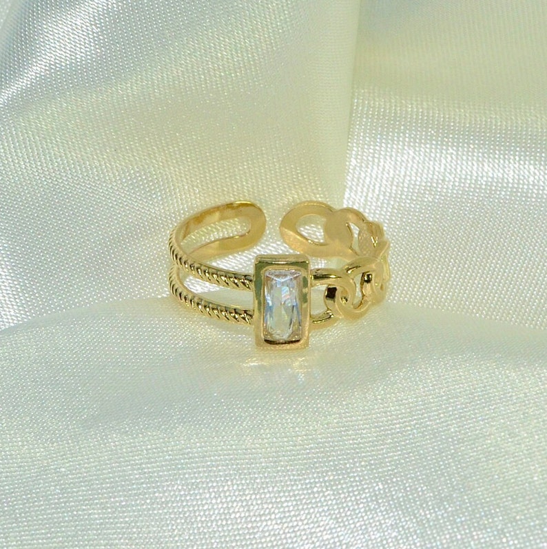 May include: Gold ring with a clear rectangular stone set in a gold frame. The ring has a chain-link design on one side and a twisted rope design on the other.