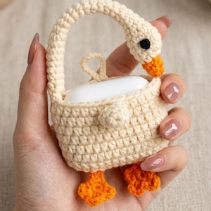 May include: Crocheted AirPods case in the shape of a goose. The case is cream-colored with orange feet and beak, and a black eye. The handle is arched, and the case is held in a hand.