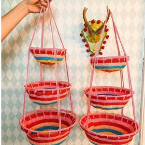 May include: Two hanging crochet baskets with three tiers each. The baskets are primarily red with rainbow stripes and pink rope hangers. The background features a wall with a diamond pattern and a decorative deer head.