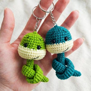 May include: Two handmade crochet keychains. One is green, the other is teal. Each keychain features a smiling face with black eyes and a cream-colored band. The bottom of each keychain is knotted. The keychains are attached to silver key rings.