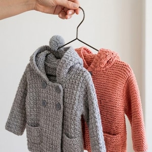 Puede incluir: Dos chaquetas con capucha de manga larga hechas de hilo de punto grueso, una en gris jaspeado y otra en rosa coral apagado. La chaqueta gris presenta un cierre de doble botonadura, dos bolsillos delanteros y un pequeño pompón en la capucha.