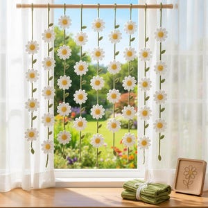 May include: A decorative daisy curtain hangs in front of a window. The curtain features white crocheted daisies with yellow centers and green stems. The window looks out onto a green garden. A wooden rod holds the curtain.