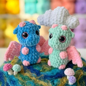 Op de afbeelding: Twee schattige gehaakte draken, één blauw en één mintgroen, met roze accenten en vleugels, zitten op een kleurrijke, getextureerde basis. Kleine gehaakte paddenstoelen en een witte wolk maken de grillige scène compleet. Een charmant, handgemaakt ambacht.