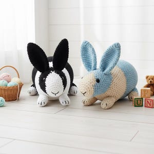 May include: Two crocheted bunny rabbits. One is black and white, the other is blue and cream. The bunnies have long ears and are lying down. A small basket of yarn and wooden blocks are in the background.