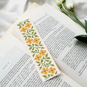 May include: A white bookmark featuring a cross-stitch design of yellow flowers and green leaves. The bookmark is positioned on top of an open book.