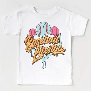 May include: White t-shirt featuring a baseball-themed graphic. The design includes a light blue baseball with red stitching and sprinkles, flanked by two pink ice cream scoops. The words "Baseball Lifestyle" are written in an orange, dripping font.