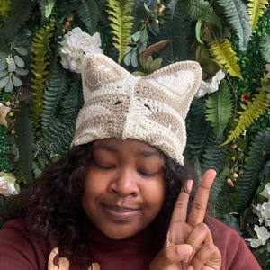 May include: A cream and tan crocheted cat ear hat with a striped pattern and pointed ears. The hat is worn by a person in front of a wall of green foliage and white flowers. The person is wearing a maroon sweatshirt with the word "Men" on it.
