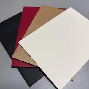 May include: A stack of blank cards in various colors: black, red, tan, and white. The cards are arranged in a slightly overlapping fan shape, showcasing their different hues and textures. The cards are likely for crafting or stationery purposes.