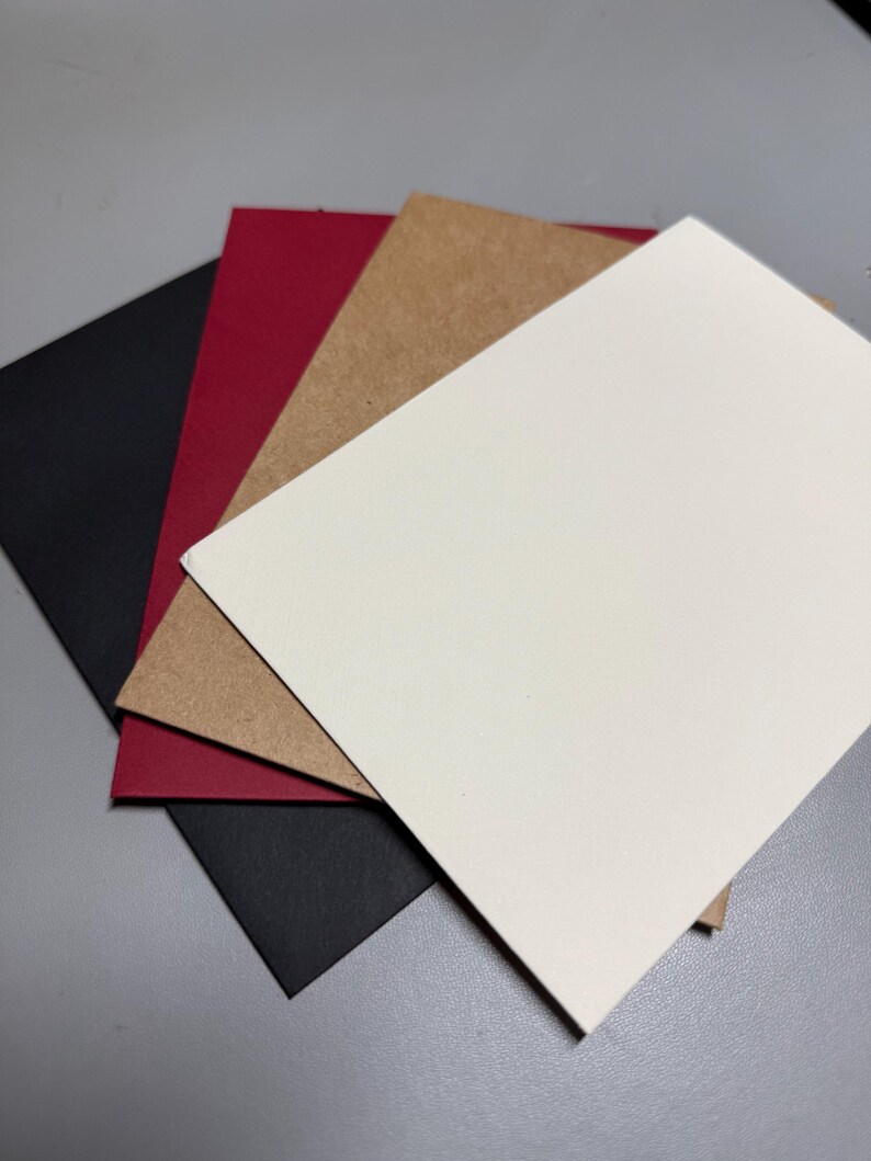 May include: A stack of blank cards in various colors: black, red, tan, and white. The cards are arranged in a slightly overlapping fan shape, showcasing their different hues and textures. The cards are likely for crafting or stationery purposes.