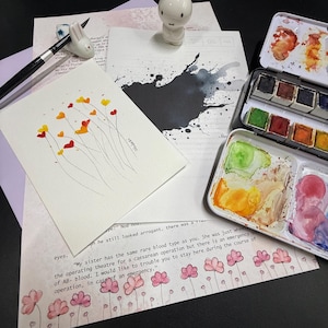 May include: A still life arrangement featuring art supplies and decorative items. Includes a watercolor painting with heart-shaped flowers, a ceramic bunny figurine, paint palettes, and a paintbrush. The scene is set on a dark surface.