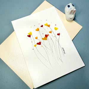May include: A watercolor painting on white paper features red, yellow, and orange heart-shaped flowers with thin black stems. A small ceramic bunny figurine sits nearby. The painting is on a cream-colored envelope, all on a blue surface.